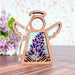 The Texas Bluebonnet Ornament by Forged Flare® is a 3.5-inch wooden angel figurine from the Mother's Angels® collection, featuring cutout wings and a halo. The angel's body showcases a stained glass–style insert depicting a Texas bluebonnet flower. This Christmas tree ornament includes a string for hanging and is set against a backdrop of lavender flowers and a light, speckled wall.