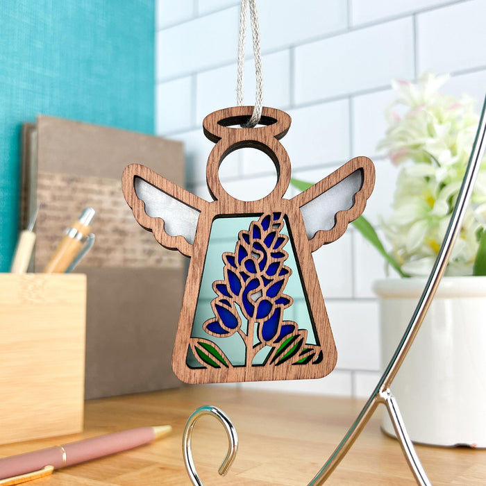 A delightful Texas Bluebonnet Ornament from Forged Flare®, part of the Mother's Angels® collection, graces a tiled backdrop. This 3.5" angel figurine features intricately designed wings and halo, with a central motif of blue flowers and wooden edges. Surrounding the scene are desk accessories and a potted plant, evoking a cozy home or office atmosphere.