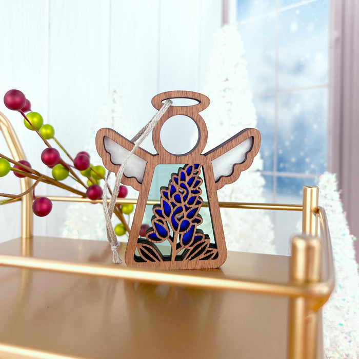 A Texas Bluebonnet Ornament from the Mother's Angels® collection by Forged Flare®, featuring white wings with a halo, stands on a gold shelf. This 3.5" wooden angel figurine is adorned with a Texas bluebonnet in the center. Perfect as flower decor, it is set against white trees, a window, and a branch with red and green berries in the background.