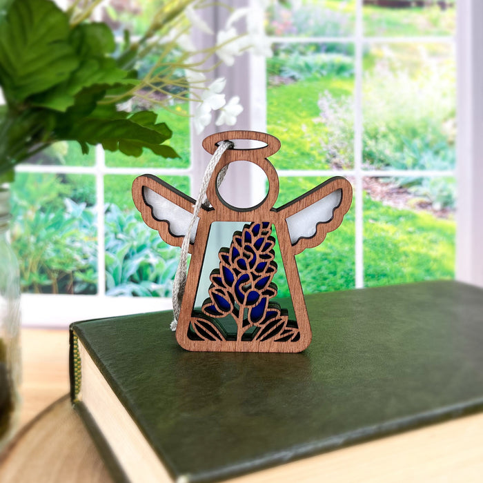 A Texas Bluebonnet Ornament | 3.5" Angel Figurine from the Mother's Angels® collection by Forged Flare® stands on a closed green book in front of a large window overlooking a lush garden. This delicate decorative piece, designed both as an angel figurine and Christmas tree ornament, features white wings, a halo, and a stained glass–style bluebonnet. A nearby vase holds green leaves.