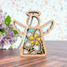 A small wooden Mother's Angels® 3.5" angel figurine rests on a wooden surface with a light floral arrangement, including white plumeria, in the background. The angel ornament from Forged Flare® features intricate cut-out designs such as a central yellow flower with green leaves and outstretched wings. It is adorned with a string for hanging.