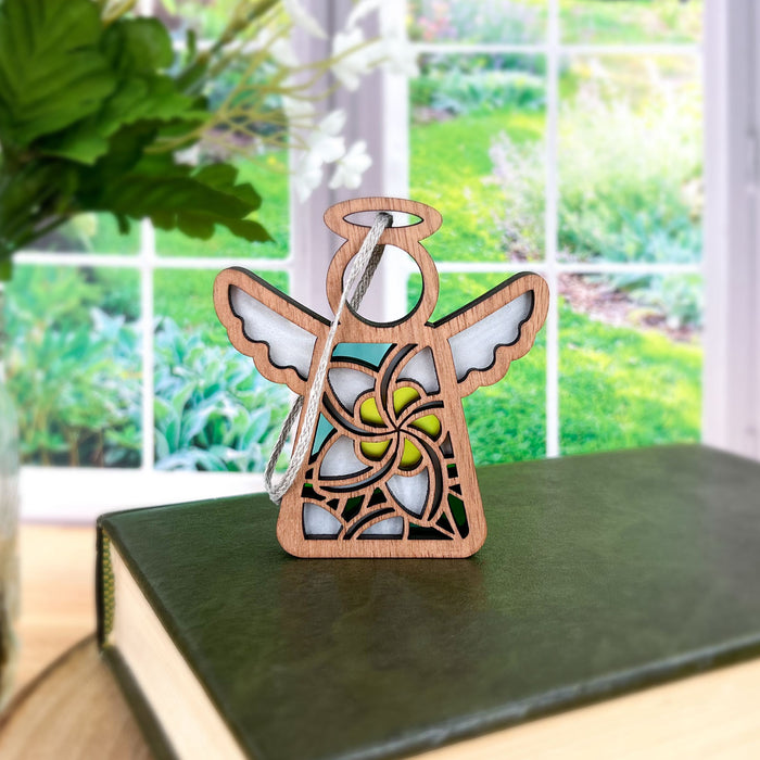 A White Plumeria Ornament 3.5" Angel Figurine from Mother's Angels® by Forged Flare® stands on a closed, green book. The angel has a halo and wings, intricately adorned with a stained glass–style plumeria design. In the background, there's a window showcasing a lush garden with greenery and flowers—a perfect setting for this elegant Hawaiian Christmas ornament.