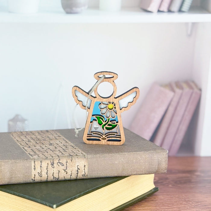 A Mother's Angels® 3.5" angel figurine by Forged Flare® stands elegantly on two closed books, resembling a treasured book ornament on a wooden table. In the blurred background, shelves brimming with more books and a small figurine are visible, making it an ideal gift for authors or book clubs.