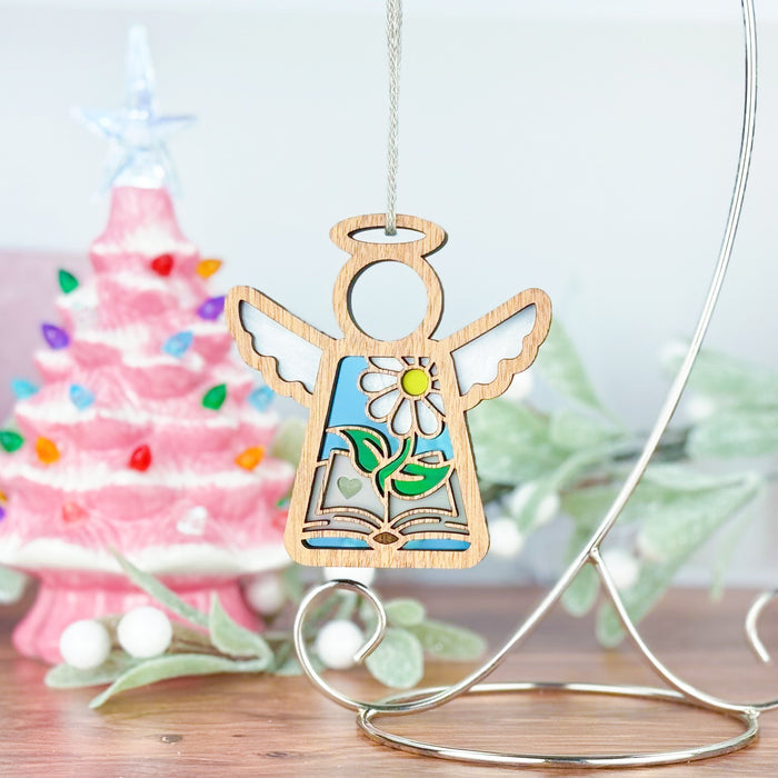 A 3.5" wooden angel figurine from the Mother's Angels® collection by Forged Flare® hangs on a stand, showcasing a floral design adorned with gemstones. Behind it stands a pink Christmas tree illuminated with colorful lights and decorations. Snowy greenery surrounds the display on a wooden surface, making it an ideal gift for book lovers or a charming addition to any book club gathering.