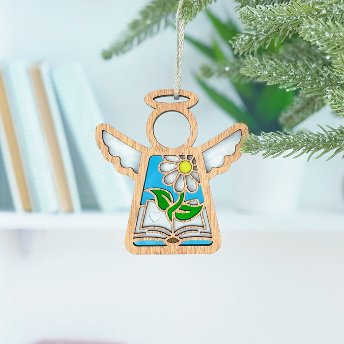 Introducing the Book Lovers Ornament—a 3.5" angel figurine from Forged Flare®'s Mother's Angels® collection. This charming wooden ornament features a stained glass design with a flower and leaves, hanging gracefully from a green branch. Perfect as an enchanting book accessory, it enhances your reading nook against the backdrop of books and a white wall, making it an exquisite gift for any book club enthusiast.