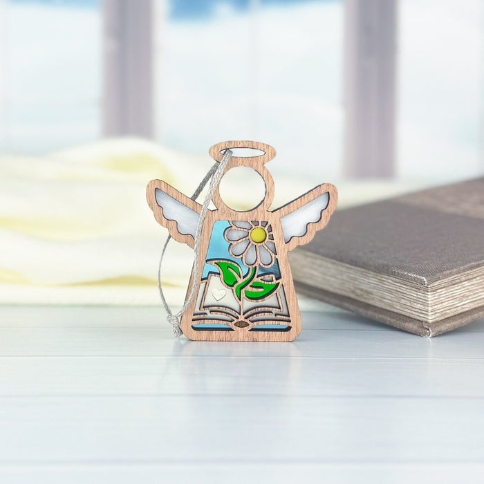 A Book Lovers Ornament from Forged Flare® stands elegantly on a table, featuring a 3.5" angel figurine from the Mother's Angels® collection. The beautifully carved design includes a flower, leaves, and an open book. In the gently blurred background are a window and another closed book—ideal as a gift for authors or any literary enthusiast's collection.