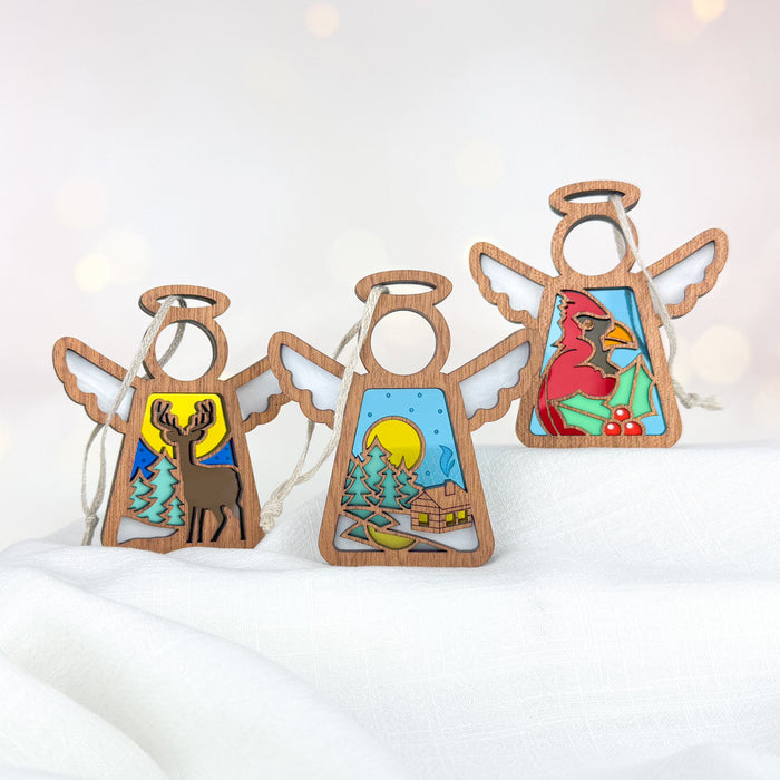 The Winter Angels 3-Piece Bundle by Forged Flare® makes for enchanting holiday decor with its wooden angel ornaments adorned with stained glass designs. One ornament, "Winter Deer," features a deer; another, "Warm Welcome," showcases a snowy cabin scene with a moon; and the third, "Winter Cardinal," displays a red cardinal on a branch. These 3.5" Mother's Angels® Christmas figurines rest gracefully on white fabric against a softly blurred background.