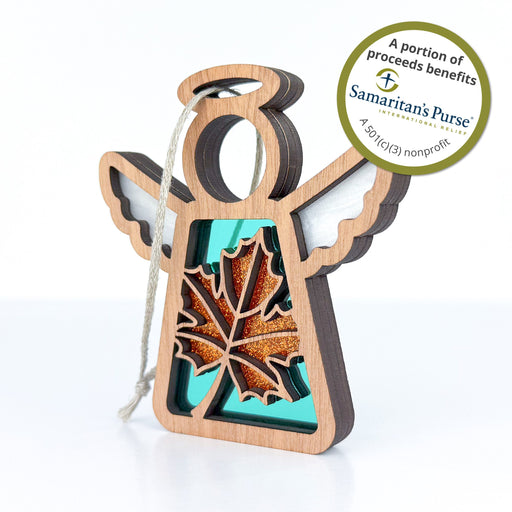 The Sugar Maple Leaf for Charity Ornament, a 3.5" angel figurine from Mother's Angels® by Forged Flare®, features a wooden angel with white wings and a cutout maple leaf design. A circular sticker highlights that proceeds benefit Samaritan's Purse, a 501(c)(3) nonprofit organization, ensuring your purchase makes a meaningful impact.