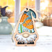 A California Poppy Ornament from Forged Flare® is displayed on a light-colored surface, showcasing a wooden gnome with a stained glass–style body adorned with orange poppies and green leaves. Blurred vases and an autumnal window view in the background make this 3.75" Nature’s Gnomes™ gift an ideal addition to your collection.