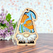 Part of the Nature's Gnomes™ collection by Forged Flare®, the California Poppy Ornament is a 3.75" wooden garden gnome featuring a stained glass–style design with orange flowers evocative of a California poppy garden, set against a blue background. The gnome, adorned with a white beard and standing on a wooden surface, is complemented by a bouquet of purple flowers and green leaves in the background.