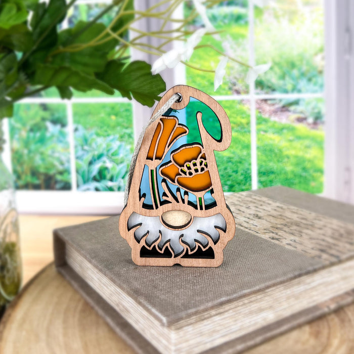 A small California Poppy Ornament from Nature's Gnomes™ by Forged Flare® is intricately carved and depicts orange poppy flowers. It is placed on a book, with a window in the background showcasing a lush garden bathed in greenery and sunlight. The scene feels serene and inviting.