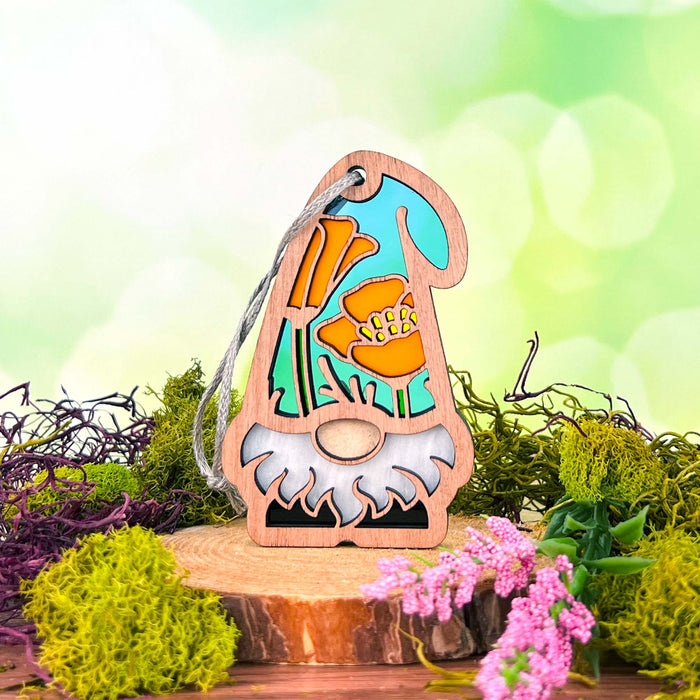 A charming, gnome-shaped ornament from Forged Flare®'s Nature's Gnomes™ collection, the 3.75" California Poppy Ornament stands on a wooden surface with its tall hat adorned in bright flowers resembling California poppies and a white beard. The background features a green and white bokeh effect, with moss and small pink flowers around the base.