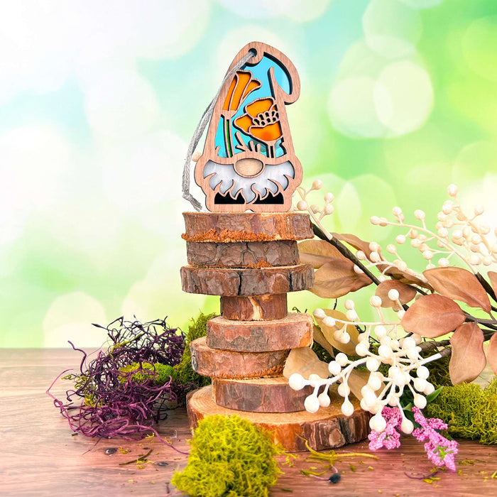 The California Poppy Ornament by Forged Flare® stands atop a stack of wooden slices, featuring a vibrant floral design. Surrounding the 3.75" garden gnome are various types of moss and dried flowers, all set against a soft, green bokeh background, creating a whimsical and natural scene. Perfect as a gift for any gardener, this charming piece from Nature's Gnomes™ collection is sure to delight.