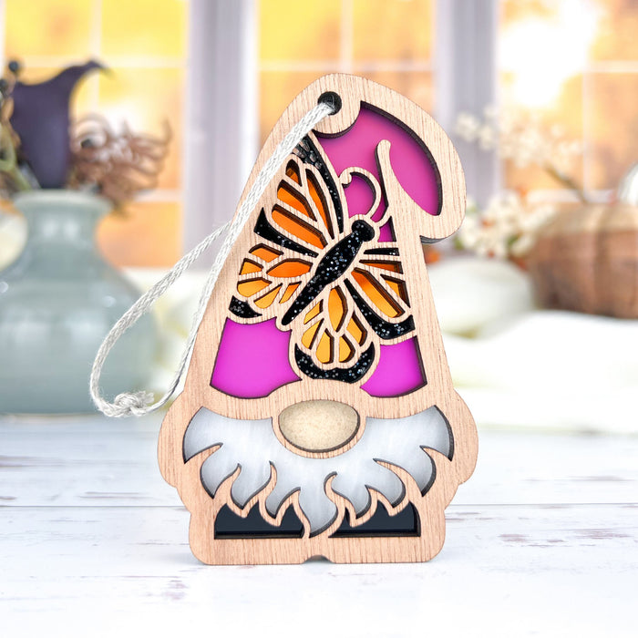 The Monarch Butterfly Ornament from Forged Flare®, part of the Nature's Gnomes™ collection, features a charming 3.75" garden gnome with a long white beard and a purple hat embellished with an intricate monarch butterfly design. The cozy indoor setting is highlighted by soft-focus vases and autumnal decor in the background, and the gnome's hat includes a string loop for easy hanging.