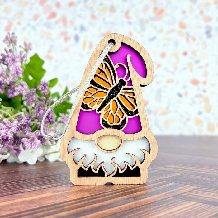 Part of the Nature’s Gnomes™ collection by Forged Flare®, this 3.75" Monarch Butterfly Ornament features a wooden garden gnome with a detailed purple hat adorned with a monarch butterfly design. The smooth finish and delicate cutouts are beautifully complemented by a background of small purple flowers and green leaves on a wooden surface.