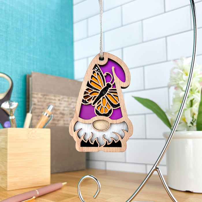 A beautiful Monarch Butterfly Ornament from Forged Flare's Nature's Gnomes™ collection, measuring 3.75 inches and featuring a gnome with a white beard and a hat decorated with a monarch butterfly design, hangs gracefully from a stand. The scene is set against a purple background, complemented by a turquoise wall, white tile, and a wooden desk adorned with various stationery items and a potted plant.