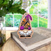 A small wooden Monarch Butterfly Ornament from the Nature’s Gnomes™ collection by Forged Flare®, featuring a purple hat adorned with a monarch butterfly, rests on a stack of books. The gnome, boasting a white beard and vibrant butterfly, is positioned indoors with the sunny garden visible through the window.