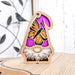 A Monarch Butterfly Ornament from Forged Flare®'s Nature’s Gnomes™ collection stands on a table, showcasing a bright pink hat adorned with a detailed monarch butterfly design. The 3.75" garden gnome ornament features a white beard and includes a string for hanging. The background reveals a blurred indoor setting with a decorative metal wall piece in view.