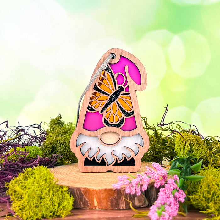 A Monarch Butterfly Ornament from the Nature's Gnomes™ collection by Forged Flare® features a 3.75" wooden garden gnome with a pink hat and white beard, standing on a wood slice base. The gnome is adorned with vibrant green moss and pink flowers, and boasts a detailed monarch butterfly design on its hat, against a blurred green background.