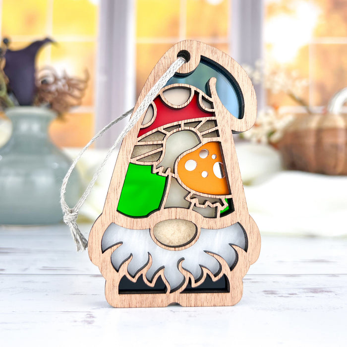 Part of the Forged Flare® Nature's Gnomes™ collection, the Toadstool Mushroom Ornament is a 3.75" decorative wooden garden gnome featuring a white beard, green shirt, and red hat adorned with toadstool mushrooms. It includes a string for easy hanging against a blurred, bright autumn scene with vases and pumpkins in the background.