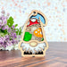 A colorful garden gnome ornament from the Nature's Gnomes™ collection by Forged Flare® stands on a wooden surface. Made of wood and stained glass, the 3.75" Toadstool Mushroom Ornament features a gnome with a red and blue hat, a white beard, and holding a toadstool mushroom. In the background, purple and white flowers with green leaves add to the charm.