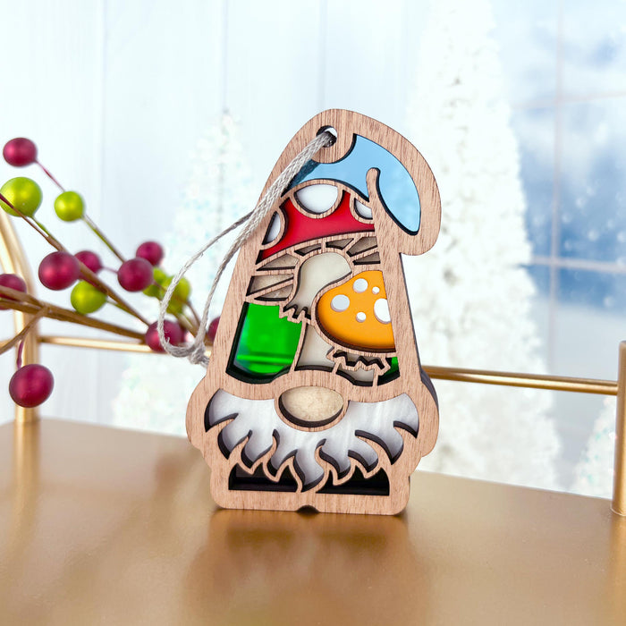 A charming Toadstool Mushroom Ornament from Forged Flare®'s Nature's Gnomes™ collection, featuring a vibrant stained glass-style design with colors like green, red, and orange, sits on a tabletop next to a sprig of red and green berries. The piece beautifully captures snowy outdoor scenery in the background.