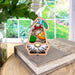 A Toadstool Mushroom Ornament from the Nature's Gnomes™ collection by Forged Flare® sits on a stack of books indoors. This 3.75" wooden garden gnome, featuring colorful stained-glass-style sections, is adorned with a green hat detailed with an orange toadstool mushroom and has a white beard. A window reveals a green garden in the background.
