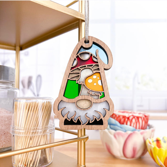 A 3.75" Toadstool Mushroom Ornament from the Nature's Gnomes™ collection by Forged Flare® hangs on a shelf. It features a garden gnome dressed in green and red, holding orange and white toadstool mushrooms. The background includes kitchen items such as toothpicks in a container, ceramic bowls, and a blurred window scene.