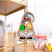 A 3.75" Toadstool Mushroom Ornament from the Nature's Gnomes™ collection by Forged Flare® hangs on a shelf. It features a garden gnome dressed in green and red, holding orange and white toadstool mushrooms. The background includes kitchen items such as toothpicks in a container, ceramic bowls, and a blurred window scene.