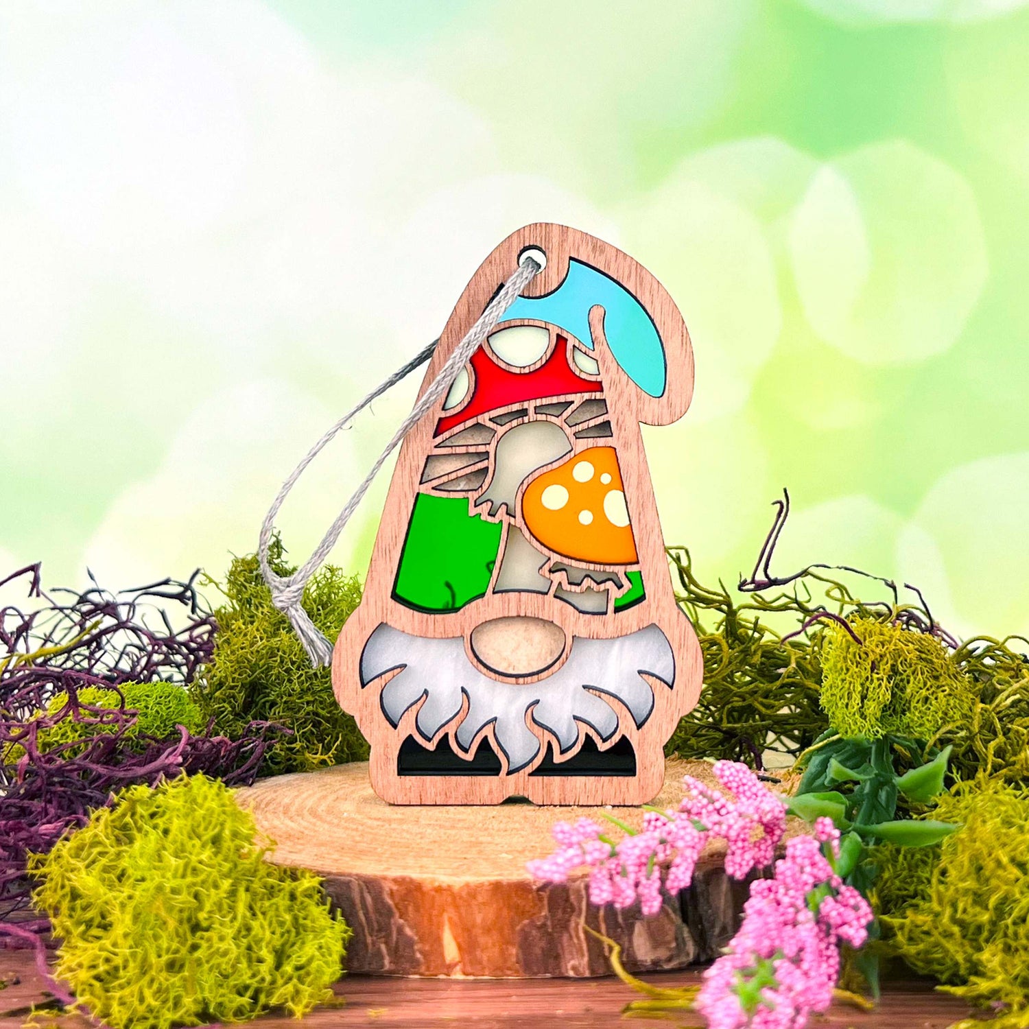 A Toadstool Mushroom Ornament from the Nature’s Gnomes™ collection by Forged Flare® stands on a rustic wooden slab, surrounded by vibrant green moss and small pink flowers. A whimsical garden gnome gift measuring 3.75" accents the scene, and the background features a blurred, soft green with light bokeh, creating a nature-inspired ambiance.