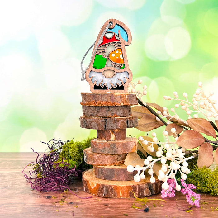 From the Nature's Gnomes™ collection by Forged Flare®, the Toadstool Mushroom Ornament is a charming 3.75" garden gnome gift. Featuring colorful stained glass with a blurred green background and bokeh lights, this delightful gnome sports a white beard, a blue hat, and holds an orange toadstool mushroom. It stands on a stack of wooden slices, surrounded by decorative foliage.