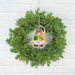 A green wreath from Forged Flare®'s Nature's Gnomes™ collection includes a 3.75" wooden ornament featuring a gnome with a toadstool mushroom cap, displayed against a white brick background.