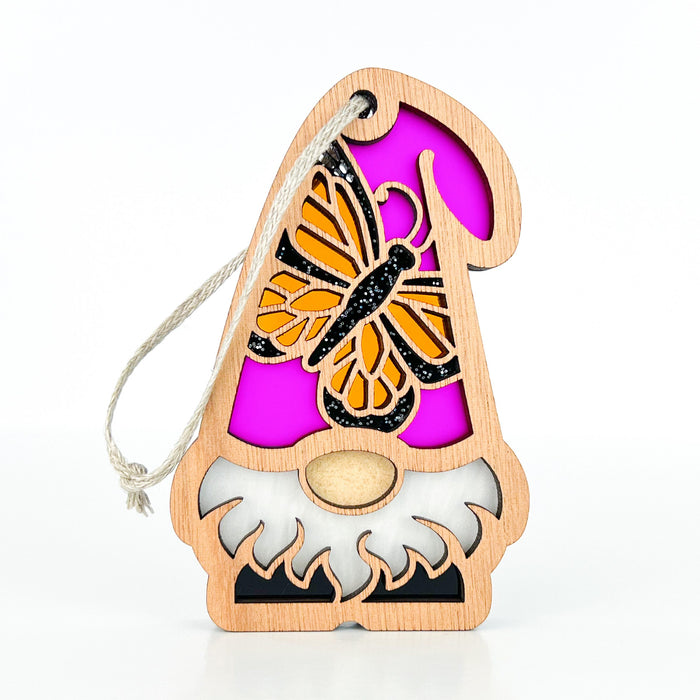 The Monarch Butterfly Ornament from the Nature's Gnomes™ collection by Forged Flare® features a wooden gnome with a cutout design adorned with a vibrant pink Monarch butterfly on its hat. The gnome has a white beard and a small nose peeking out, and comes with a natural twine loop for hanging. This 3.75" garden gnome ornament is perfect for enhancing any outdoor space.