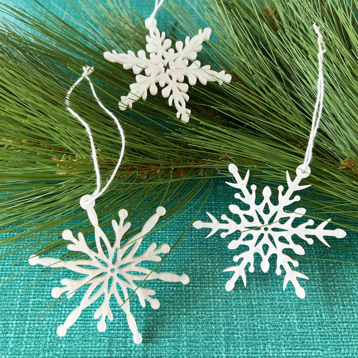 Three Wintry Wonderland Snowflakes Bundle, 3", from Forged Flare®, with unique white snowflake patterns lie on a bed of green pine needles. The background is a textured blue surface. Each snowflake, perfect as winter wonderland decorations, hangs with a white string, ready for adornment.