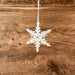 Against a textured wooden background hangs the Wintry Wonderland Snowflake - Snow Star, 3" by Forged Flare®. This delicate white ornament features detailed and intricate cut-outs, adding to its elegant appearance and making it perfect for winter decor.