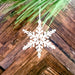 A Wintry Wonderland Snowflake - Snow Star, 3" ornament from Forged Flare® hangs from a string in front of a wooden background, near green pine needles. The white snowflake features intricate cut-out details with a symmetrical design. This unique Christmas tree ornament adds charm to your winter decor, set against the rich texture of the diagonal-patterned wood.