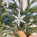 A close-up of a Wintry Wonderland Snowflake - Snow Star, 3" ornament by Forged Flare® hanging on a green Christmas tree. The intricately designed snowflake stands out against the tree's branches. The background is softly blurred, further emphasizing the festive decoration.