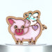 A cute, colorful pig ornament made of wood and stained glass featuring a pink body, iridescent wings, and a purple glitter heart on its rear. The pig ornament has a playful design with a whimsical smile, and it hangs from a braided linen string.