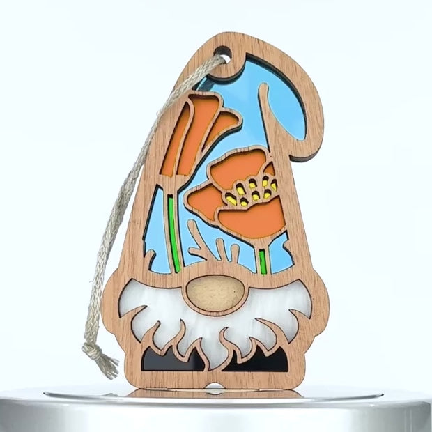 A wooden ornament shaped like a gnome features cut-out California poppies and green leaves backed by a light blue background. The gnome has a white beard and a flower hat. A white string is attached to the ornament for hanging.