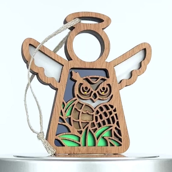 A wooden owl ornament shaped like an angel with wings and a halo, featuring a detailed engraving of an owl perched on green leaves. The ornament has a blue background and a string attached at the top for hanging.