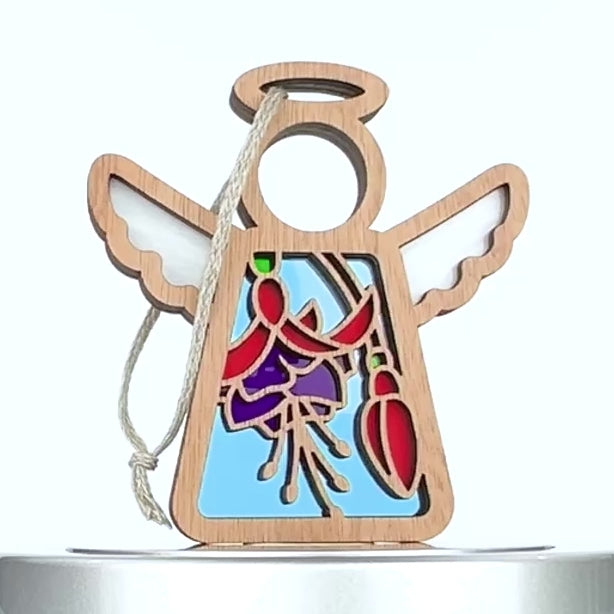 A decorative wooden ornament shaped like an angel, featuring a vibrant stained glass–style fuchsia flower design in red and purple. The angel has a braided linen string attached for hanging, making it the perfect fuchsia flower ornament for any space.