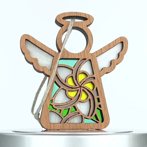 A wooden angel ornament, reminiscent of a plumeria ornament, features a circular head, winged arms, and a colorful stained glass–style body with green, blue, and yellow patterns. It has a loop of string attached for hanging against the simple white background.