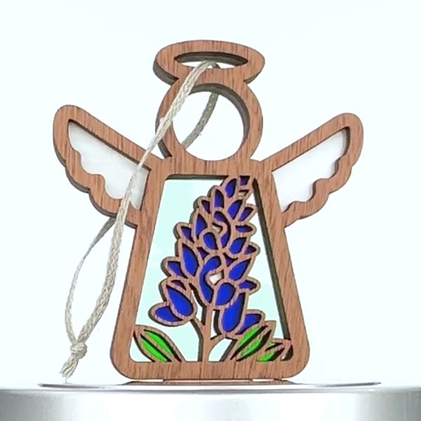 A wooden ornament shaped like an angel with wings and a halo. The center features a stained glass–style design of a bluebonnet flower with green leaves, reminiscent of Texas fields. A braided linen string is attached to the top for hanging.