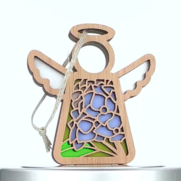 A wooden angel ornament featuring stained glass–style decorations on its body. The design includes intricate blue and purple floral patterns with green leaves, reminiscent of a hydrangea ornament. A string is attached to the top for hanging.