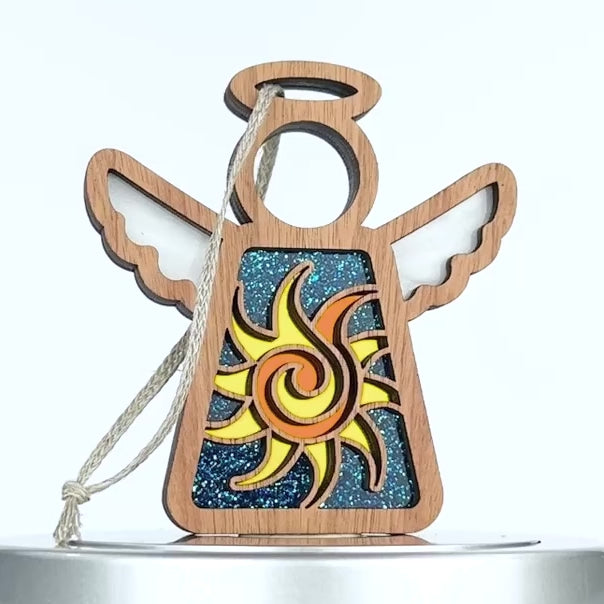 A wooden angel ornament with outstretched wings and a halo. The angel's torso features a captivating cut-out design showcasing a celestial sun against a blue glittery background. It includes a light-colored hanging string attached to the top.