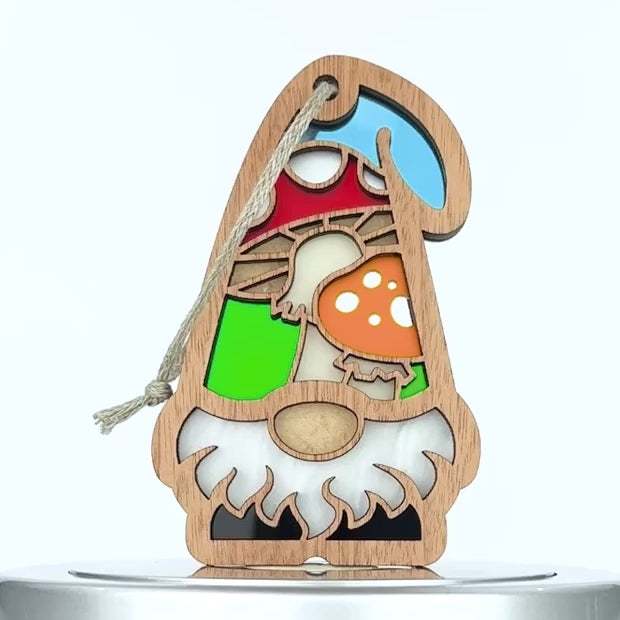 A decorative wooden gnome ornament in the shape of a gnome, featuring a colorful stained glass–style design with a red hat, a yellow toadstool mushroom, and a green patch. The gnome has a white beard and hangs by a braided linen string through a hole in the tip of the hat.