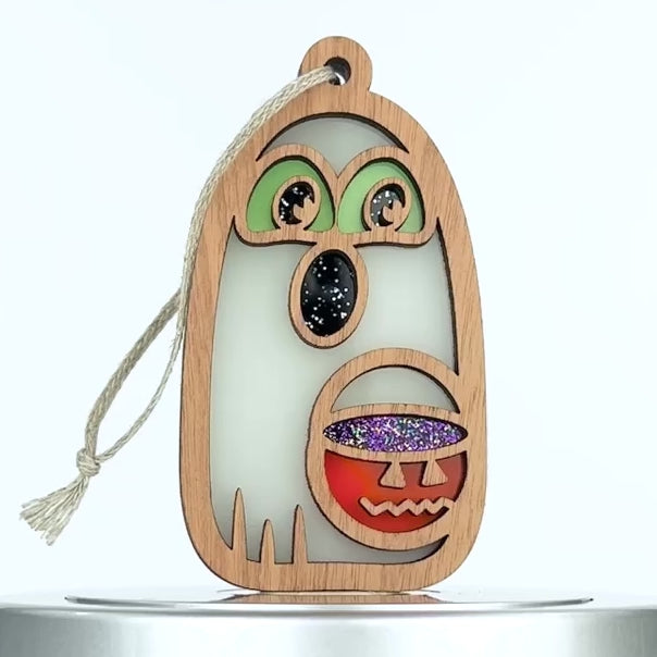 A wooden ghost ornament shaped like a specter with green eyes and an open mouth. The ghost holds a jack-o'-lantern bucket filled with glitter. A string is attached for hanging. The background is plain white.