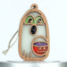 A wooden ghost ornament shaped like a specter with green eyes and an open mouth. The ghost holds a jack-o'-lantern bucket filled with glitter. A string is attached for hanging. The background is plain white.