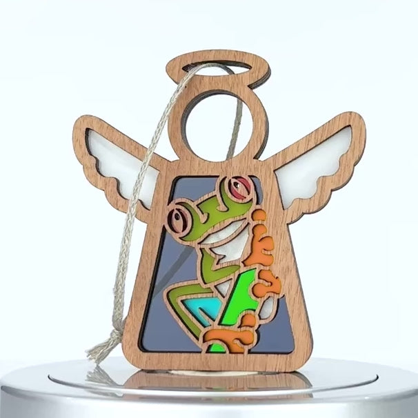 A decorative ornament made of wood depicts an angel with outstretched wings and a halo. The center features a colorful, whimsical tree frog with large eyes and a green body. A braided linen string at the top allows it to be hung as decoration.