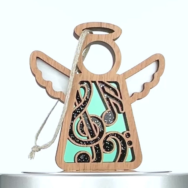 A wooden angel music ornament with a decorative cut-out design on its body, featuring intricate musical notes. The wings are white, and there's a string attached to the top for hanging. The ornament has a halo above its head and a blue backdrop behind the musical notes.